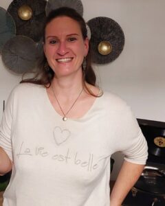 Blandine Coudurier-Dufour, jeune femme souriante car elle fête sa certification de coach de vie certifiée. Elle porte un pull sur lequel est inscrit "La vie est belle" avec un coeur au-dessus de la phrase.